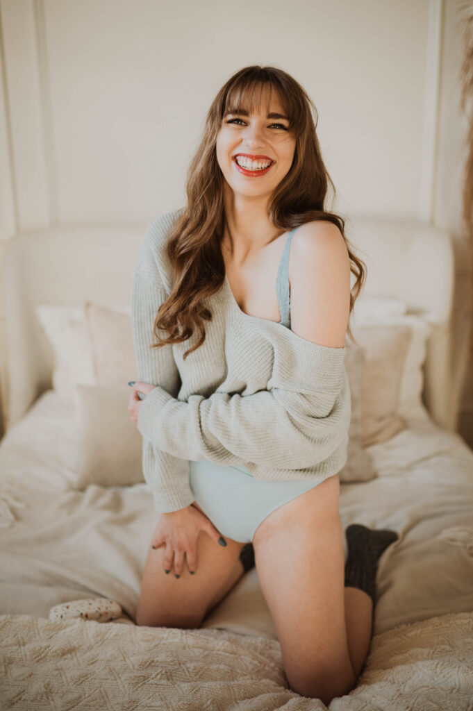 A woman smiles for the camera in a oversized sweater during a boudoir photoshoot