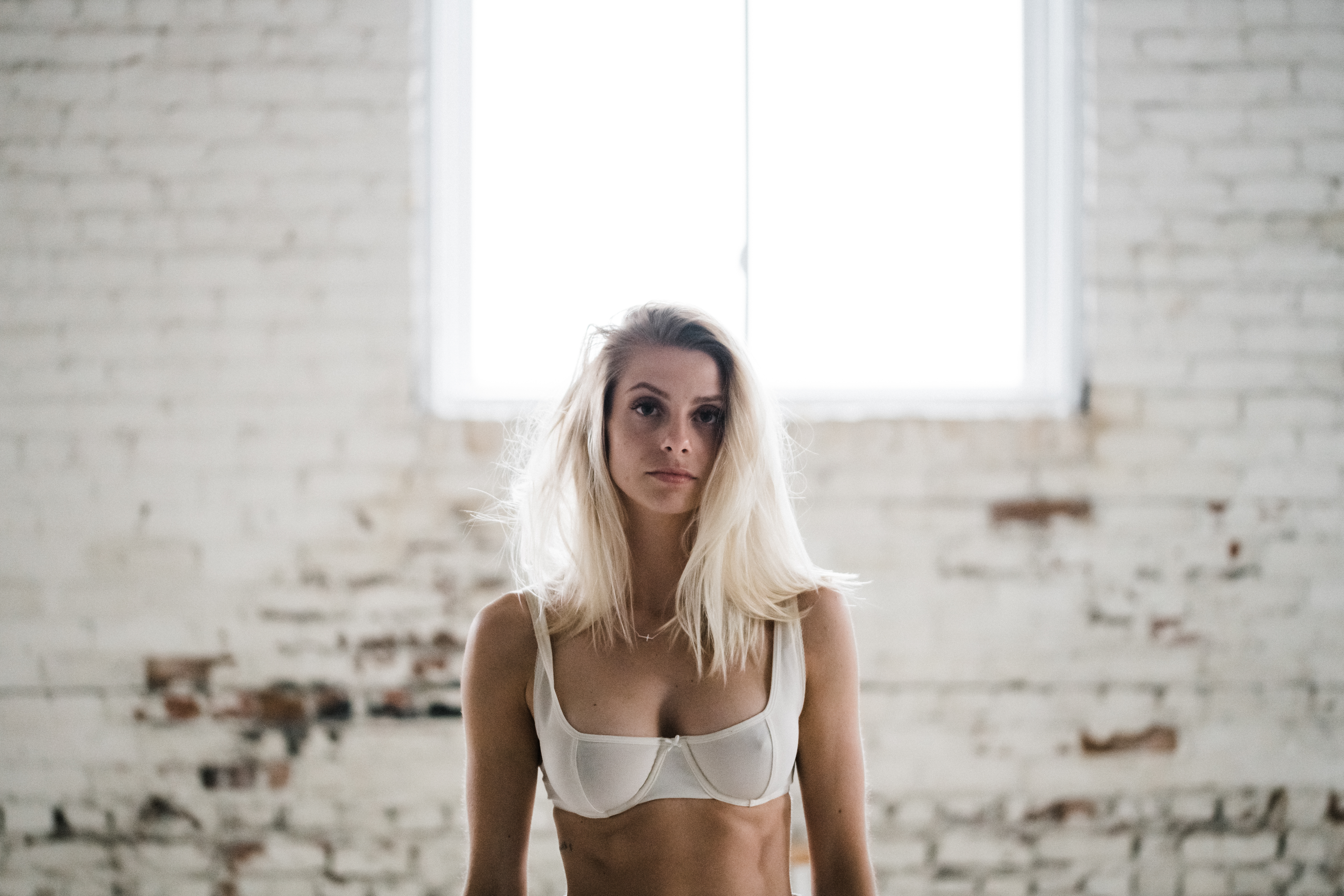 A woman stands in front of a white brick wall in jeans and a bra for a boudoir session